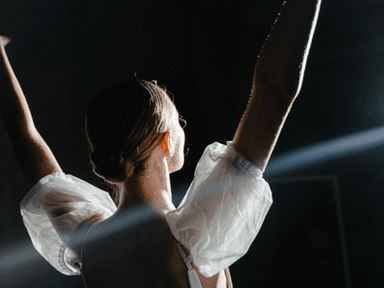 A dancer with their back turned raises arms, illuminated by spotlight in a dark space